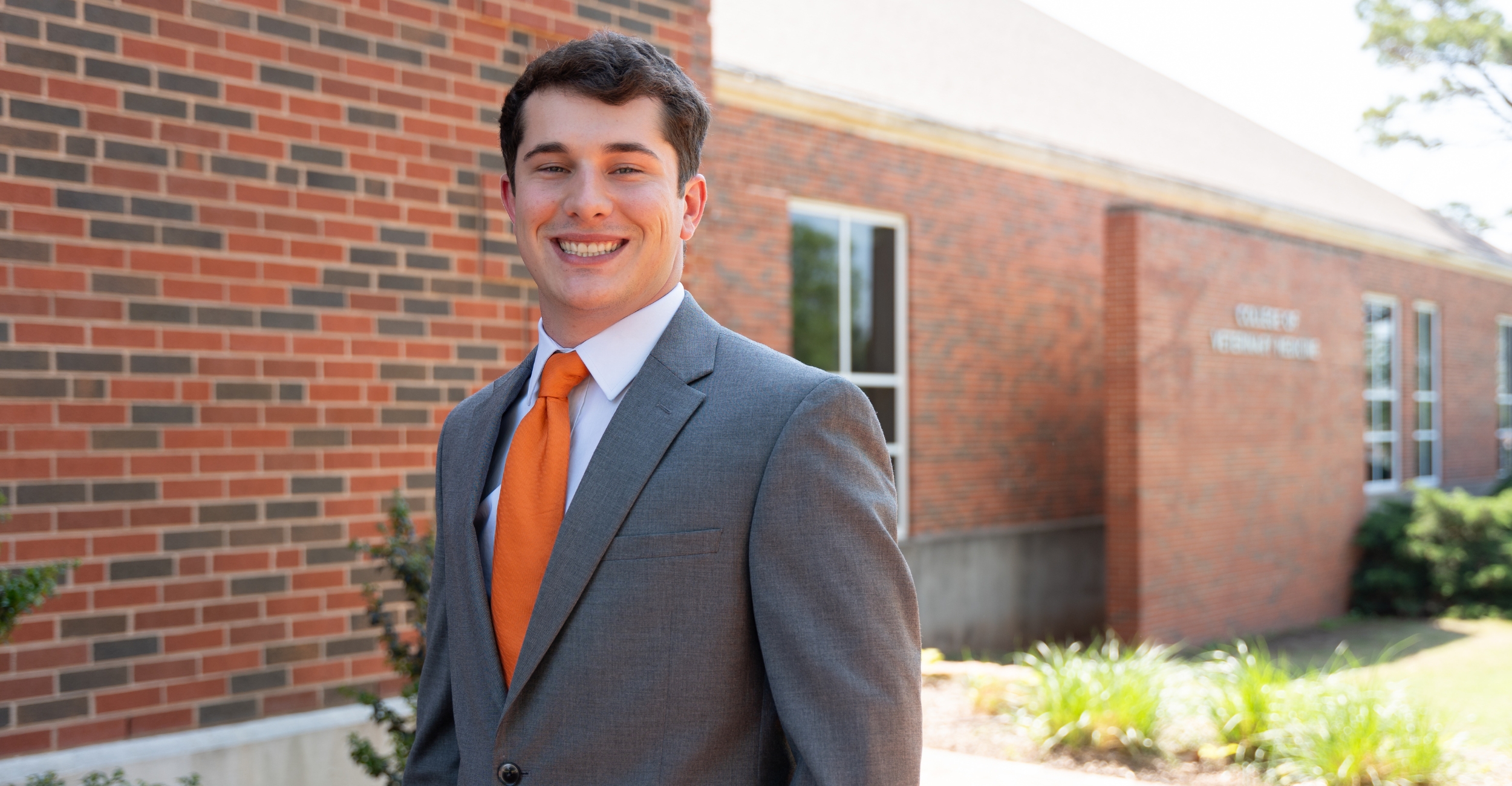 A person wearing a gray suit and orange tie stands outdoors in front of a brick academic building with signage partially visible in the background, suggesting a professional or campus portrait setting.