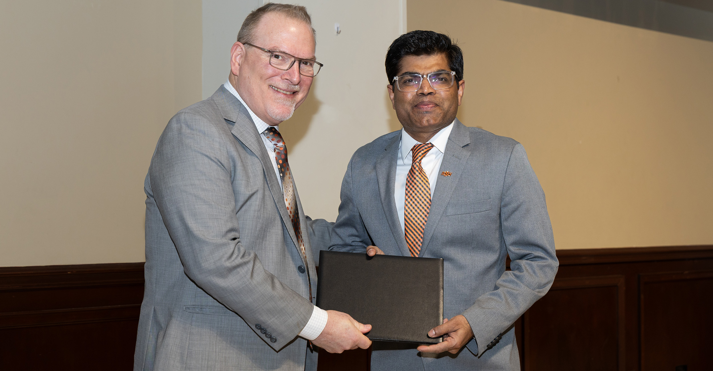 Two individuals in business attire stand indoors while jointly holding a framed certificate, indicating a formal academic or research award presentation.
