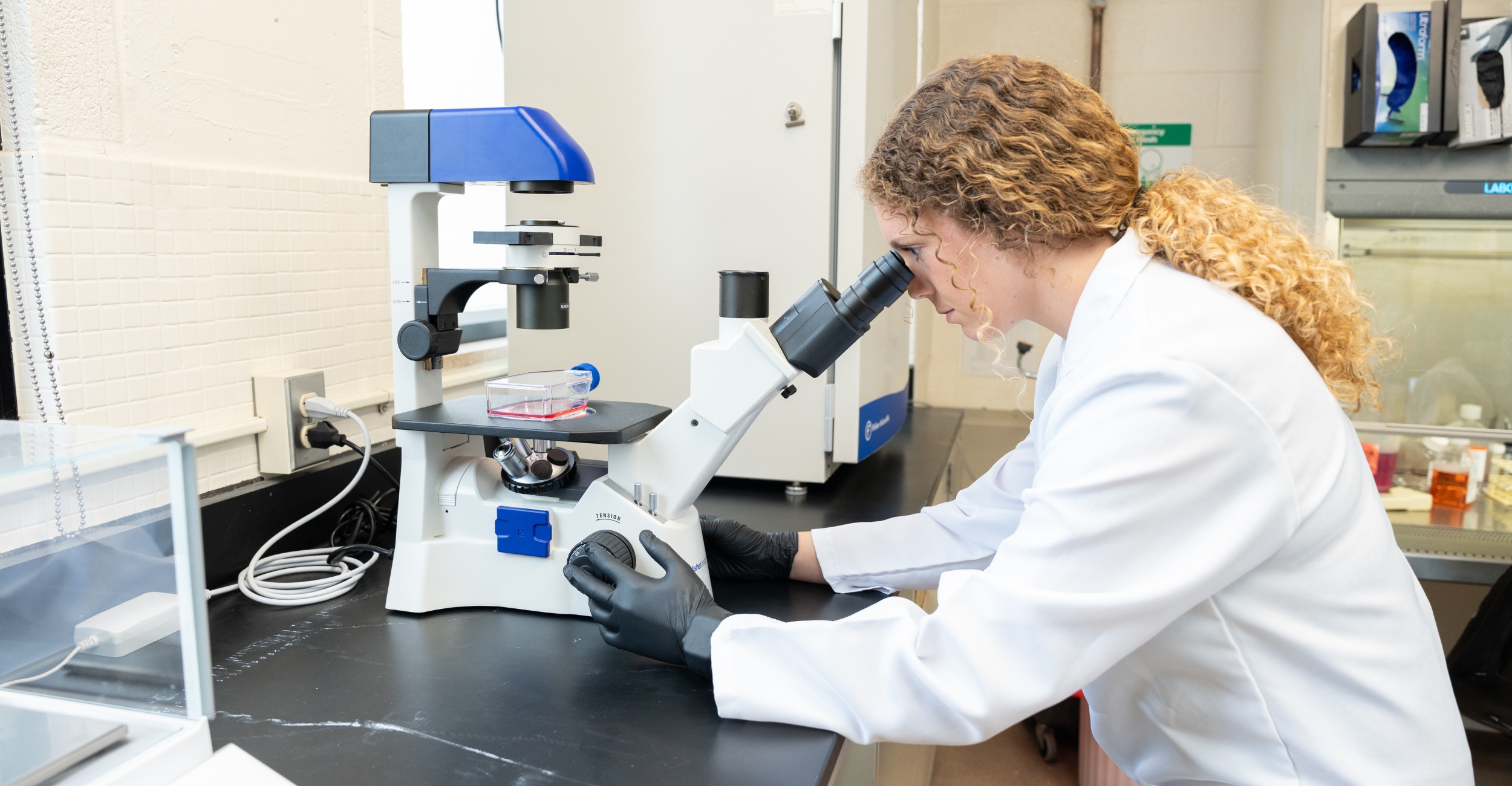 A college student in a white lab coat and lab setting looks into a microscope.