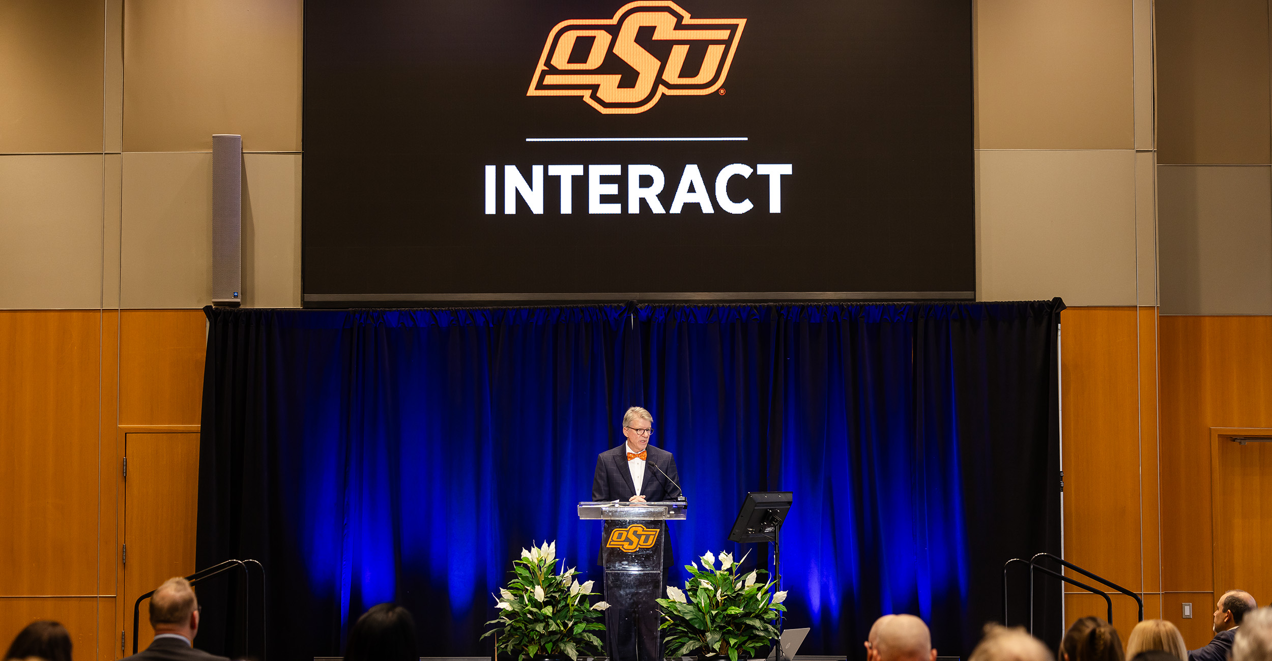 Person standing at a clear podium on a stage with blue curtains, speaking during an event. A large screen above displays the OSU logo and the word ‘INTERACT.’ Green plants decorate the front of the stage, and audience members are seated in the foreground.
