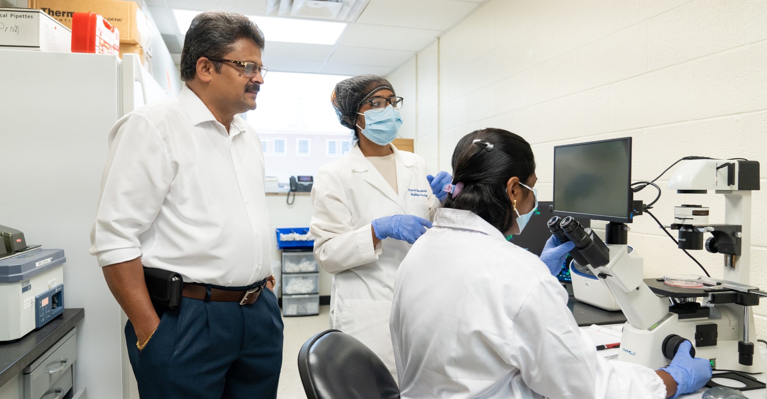 Three people wearing lab coats and protective gear work together in a laboratory, with one person using a microscope while the others observe and discuss the research process.