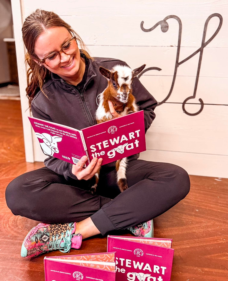 A person sits cross‑legged on a wooden floor holding a baby goat while reading a children’s book titled Stewart the Goat, with additional copies of the book displayed nearby in a cozy indoor setting.