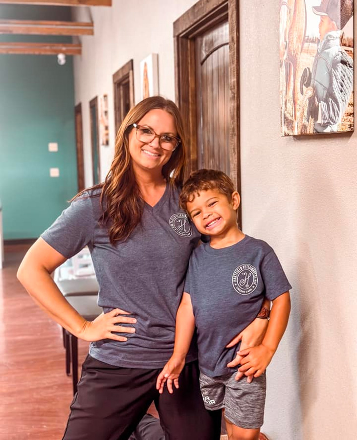 An adult and a child wearing matching T‑shirts stand together in a hallway with framed artwork on the wall, suggesting a family‑friendly or community setting.