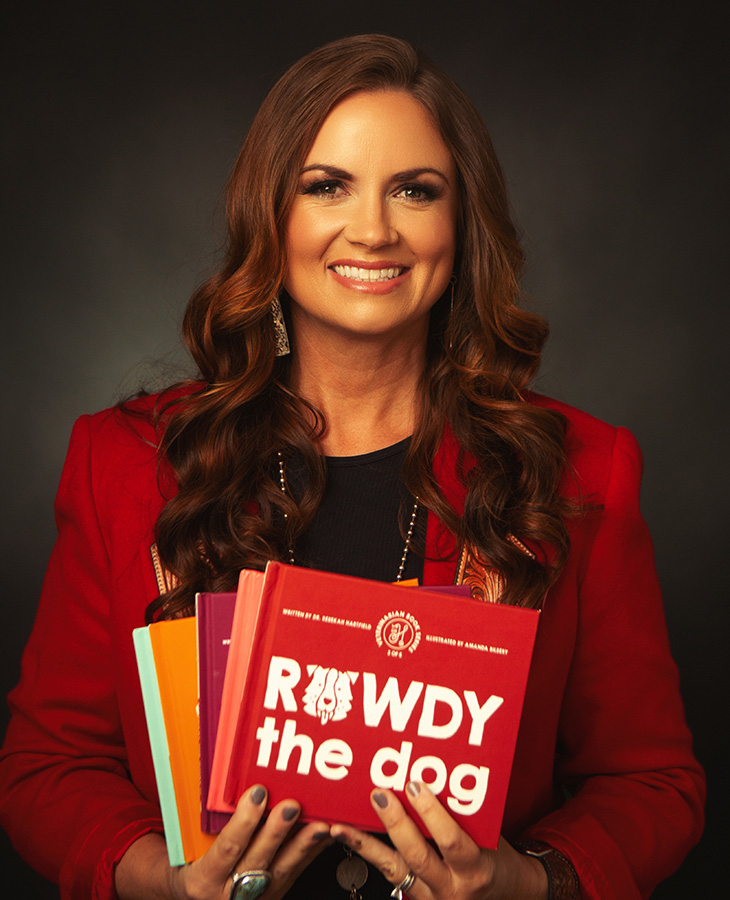 An individual wearing a red jacket stands against a dark studio background while holding a stack of children’s books titled Rowdy the Dog, suggesting a promotional or educational reading context.