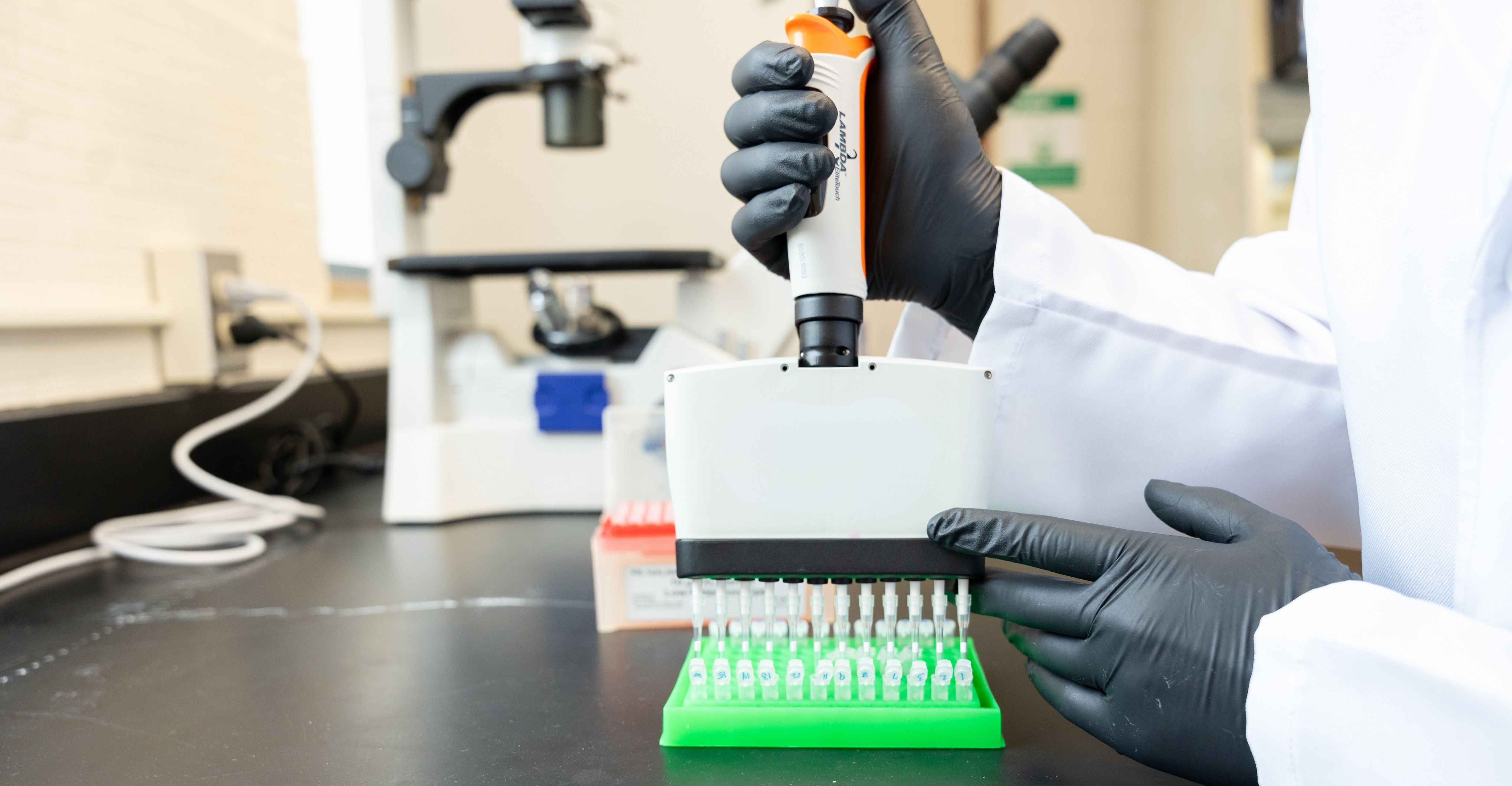 Person wearing black gloves and a white lab coat using a pipette to fill small tubes arranged in a green rack with laboratory equipment including a microscope in the background.