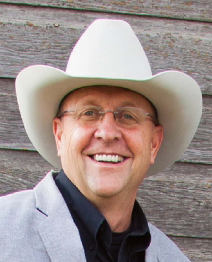 A close-up portrait shows a person wearing glasses and a light-colored cowboy hat against a wooden exterior wall, presenting a professional or promotional headshot.