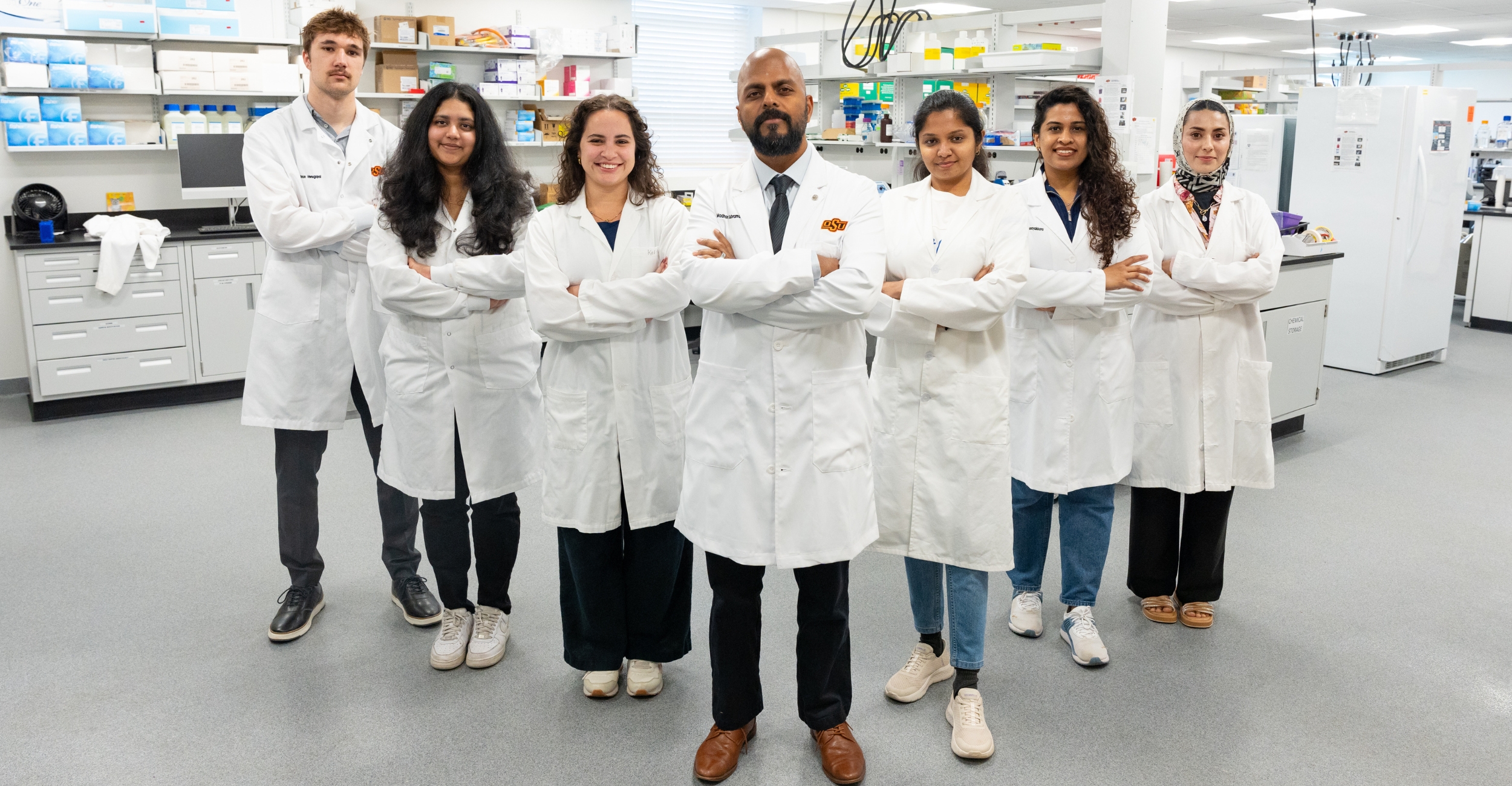 A group of seven researchers in white lab coats stands in a laboratory with arms crossed, posed in front of benches stocked with scientific equipment and supplies to convey a collaborative research team.