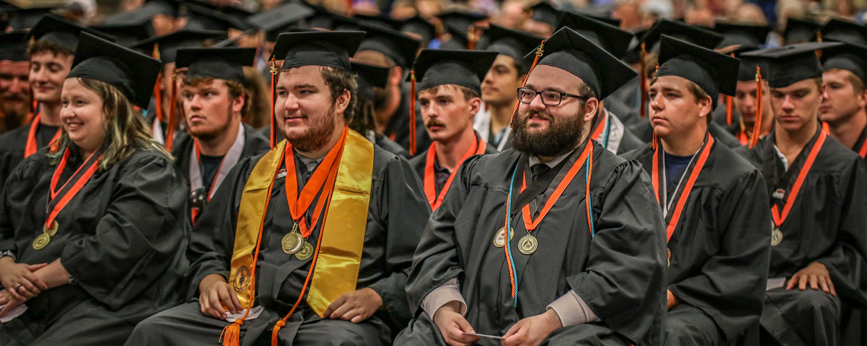 A group of OSUIT graduates sit in rows wearing black caps and gowns. Each student has a bronze graduation medallion on an orange-and-black ribbon, and some wear additional regalia, including a yellow Phi Theta Kappa stole and a turquoise first-generation honor cord.