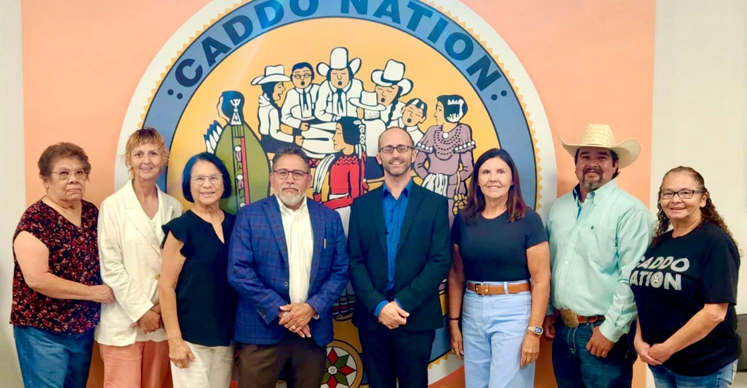 A group of eight people stands in front of a colorful mural featuring the words "Caddo Nation" and illustrations of people in traditional attire. The group includes men and women, some dressed in business attire and others in casual or traditional clothing, posing and smiling for the photo.