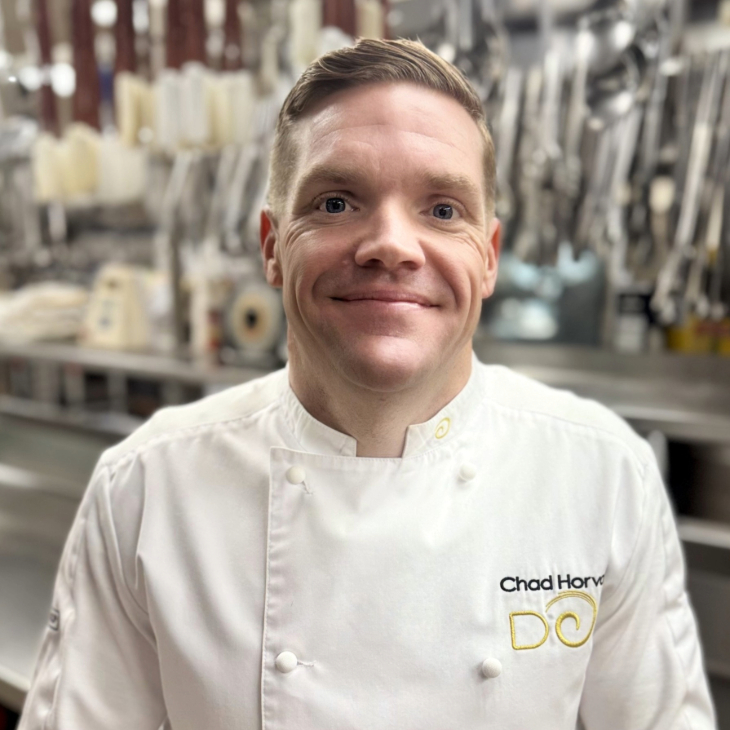 A smiling man in a white chef’s coat stands in a professional kitchen, surrounded by hanging utensils and cooking tools in the background.