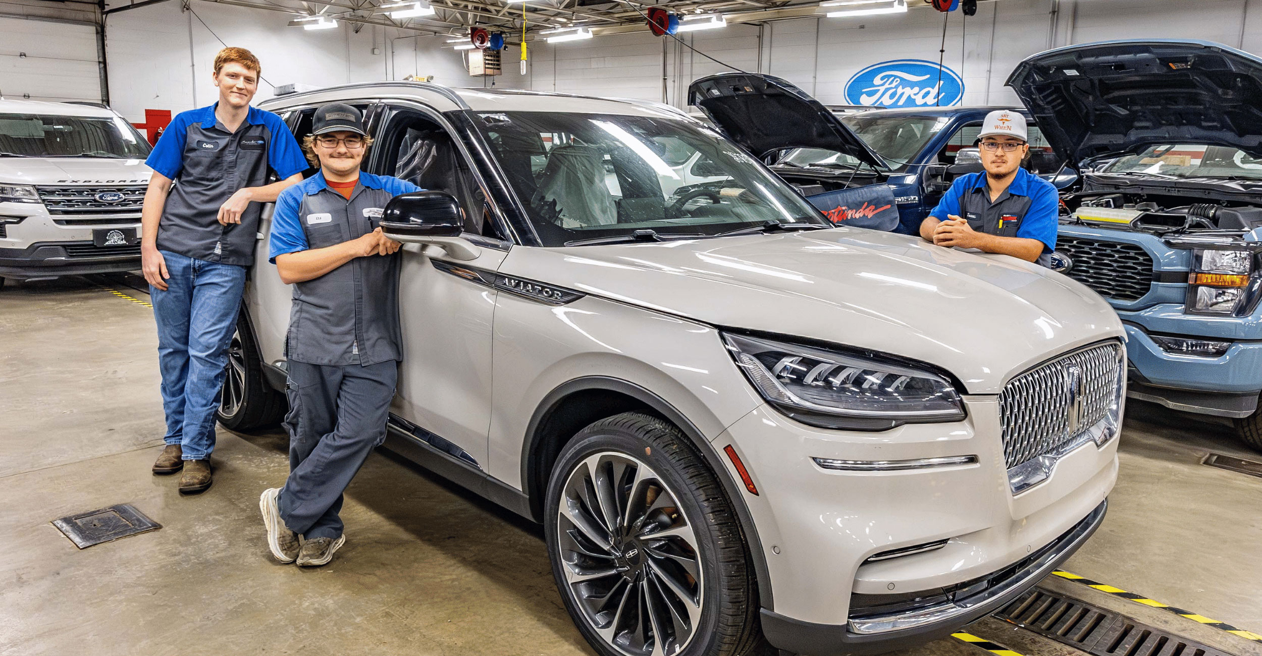 Three OSUIT Ford ASSET students pose in a campus automotive lab with a 2024 Lincoln Aviator, donated by Ford Motor Company. Other Ford vehicles and service equipment are visible in the background.
