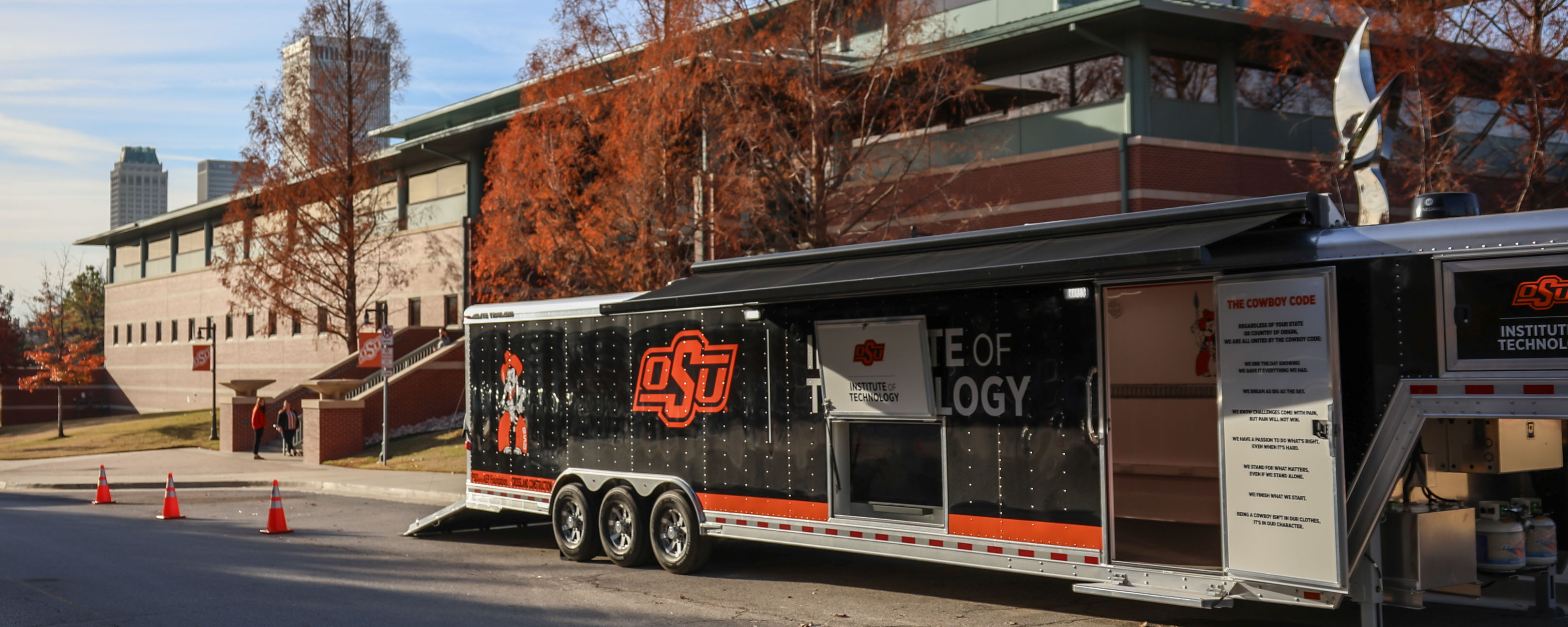 A large OSU Institute of Technology (OSUIT) branded trailer is parked on a street near a modern brick and glass building with autumn-colored trees in the background. The trailer features the OSU logo, the words 'Institute of Technology,' and 'The Cowboy Code' displayed on its side. Part of the trailer is open, revealing an interior workspace. Orange traffic cones are placed nearby. In the distance, a few people are walking on a pathway, and Tulsa's downtown skyline is visible in the background.