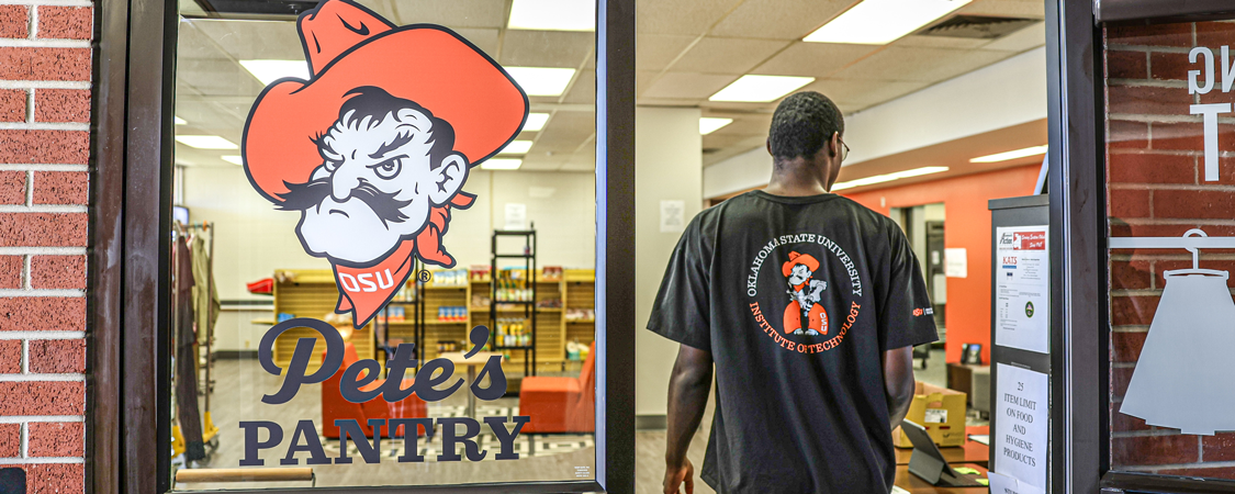 A person wearing an Oklahoma State University Institute of Technology T-shirt walks into Pete’s Pantry, seen through a glass door with the Pistol Pete logo and “Pete’s Pantry” text. Shelves stocked with food items are visible inside.