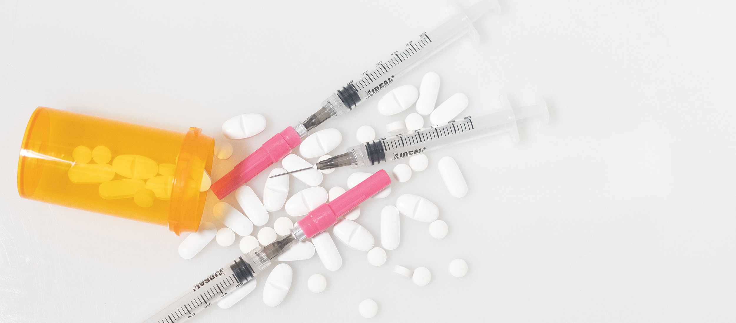 There is an orange pill bottle, numerous white pills and three syringes spread across a white surface.