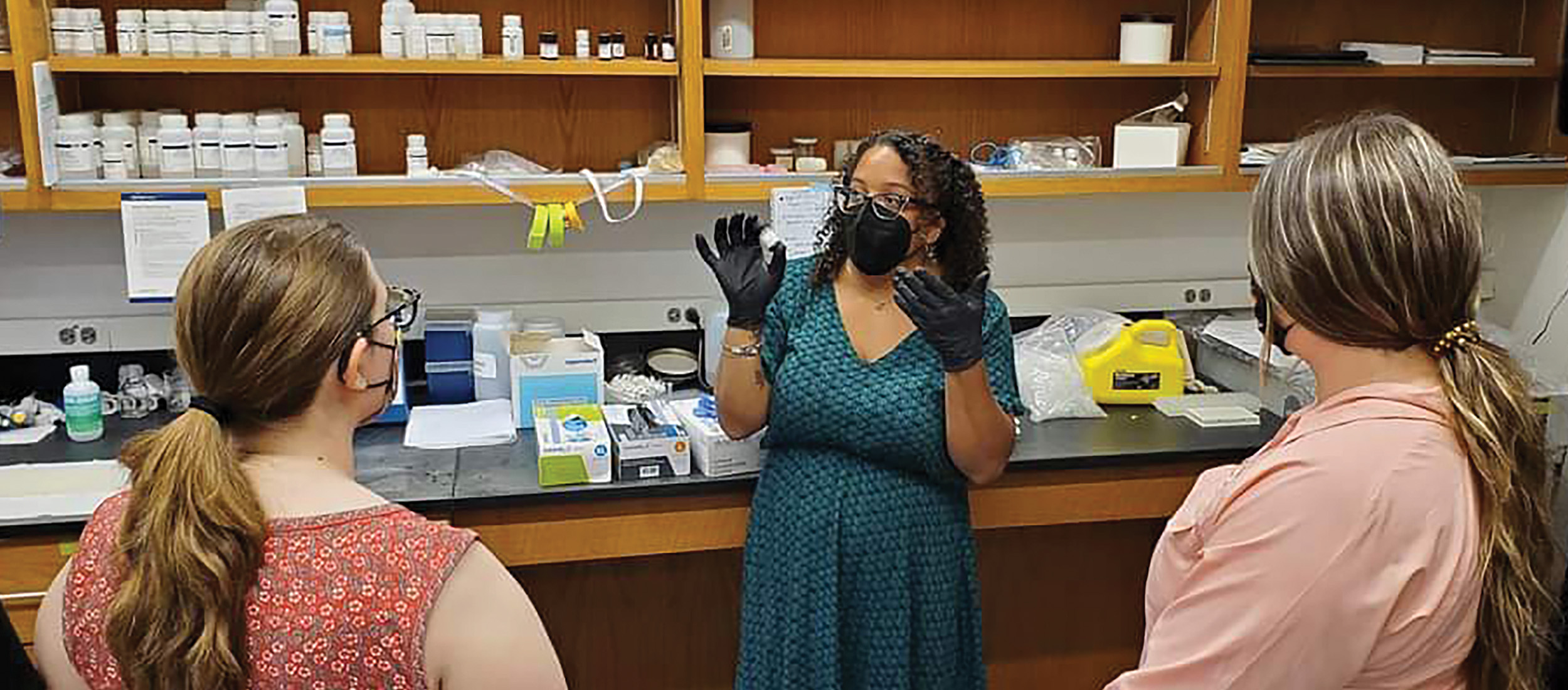 Jessica Ware (center) explains gel electrophoresis to Samantha Hittson (left) and Melissa Reed. They are in a lab room with bottles on the shelves in the background.