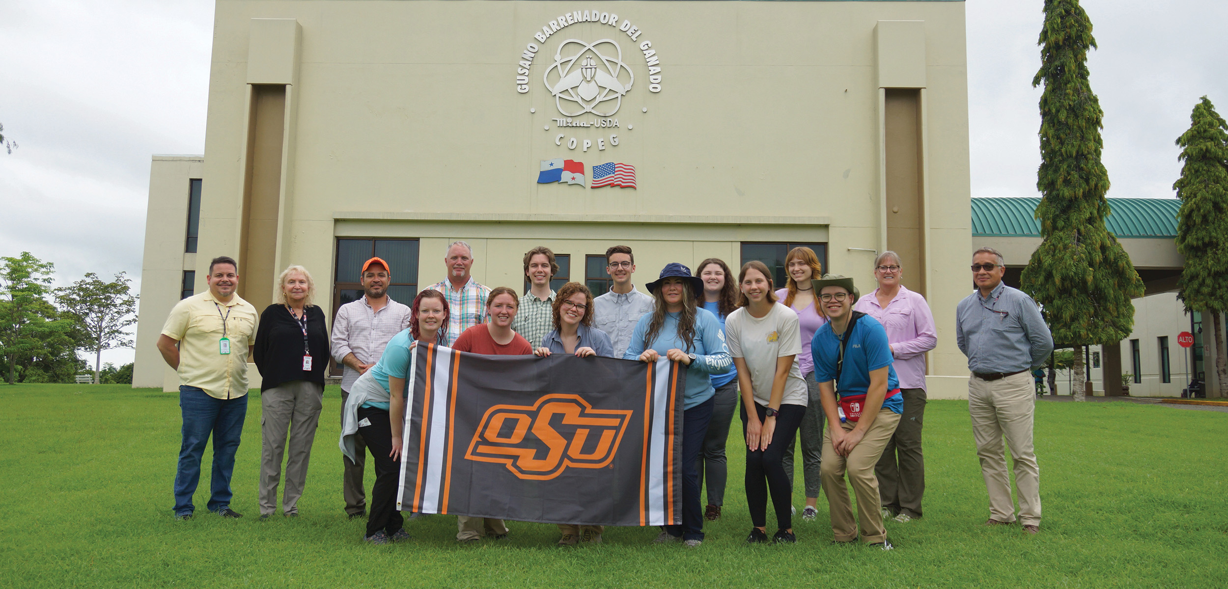 Study-abroad participants visit the COPEG lab. The students are holding an OSU flag in front of the building.