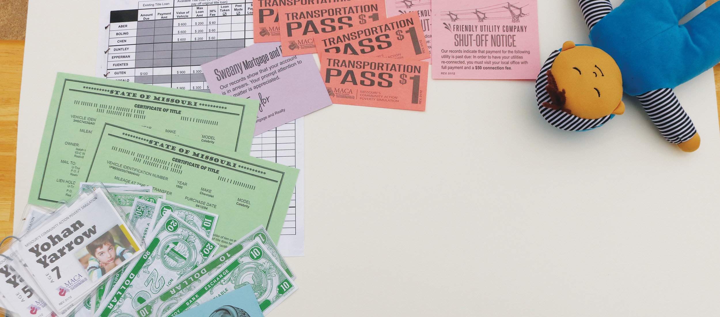 Various poverty simulation materials including fake money, certificates, a doll and more are spread across a table.