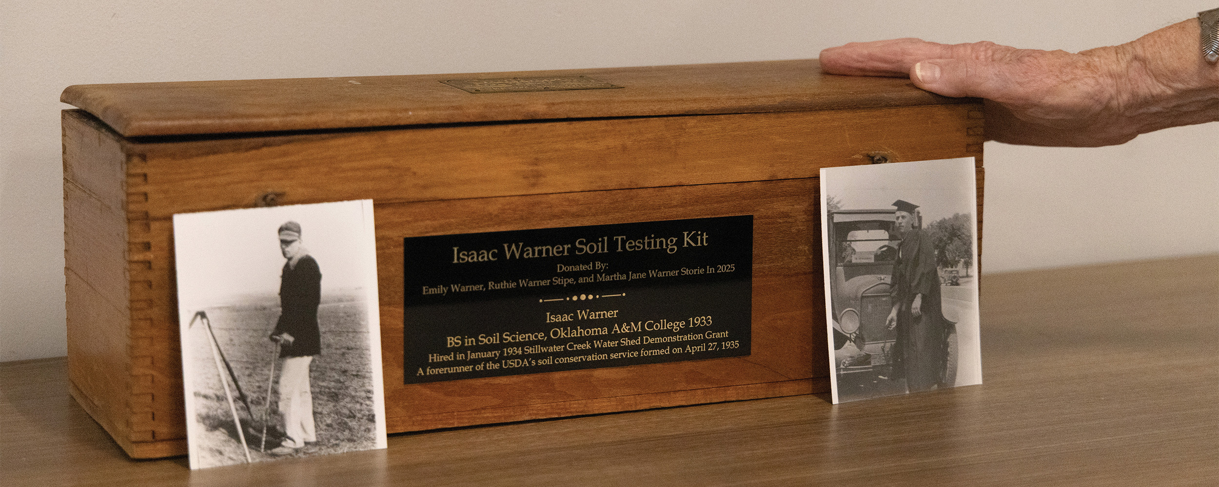 A wooden box labeled “Isaac Warner Soil Testing Kit,” displayed on a table with an engraved plaque describing its history and donation. On either side of the box sit two vintage black-and-white photographs: one showing a man standing in a field with soil sampling tools, and the other showing a man in a graduation cap and gown posing beside an earl-model car. Emily Warner rests her hand softly on the top of the box.