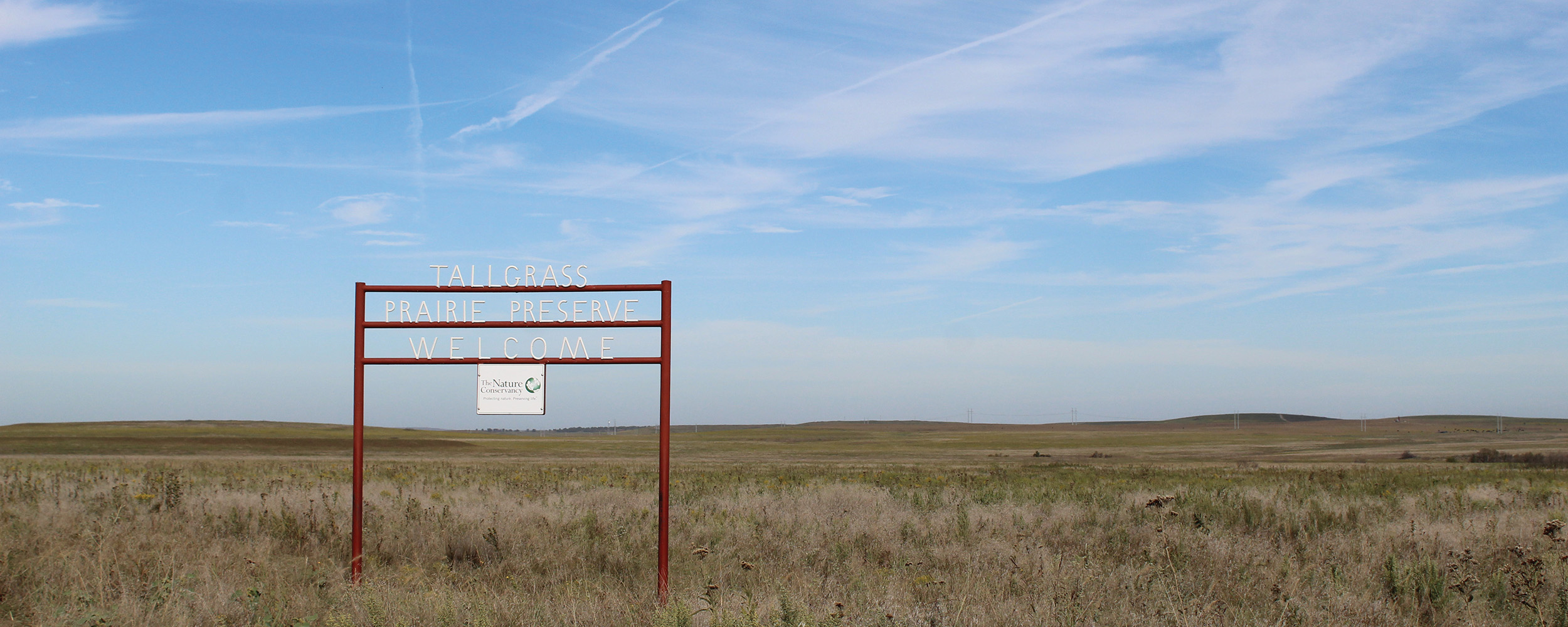 A wide view of a tallgrass prairie with a red pipe sign saying “Tallgrass Prairie Reserve Welcome” in white.