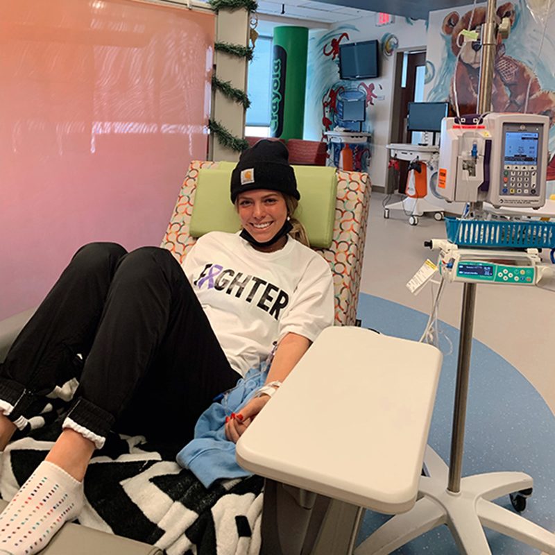 A teenage girl wears a beanie hat and sits in a hospital bed. Despite going through cancer treatment, she smiles. Kid-themed decorations, including an image of a teddy bear and giant crayon, are behind her.