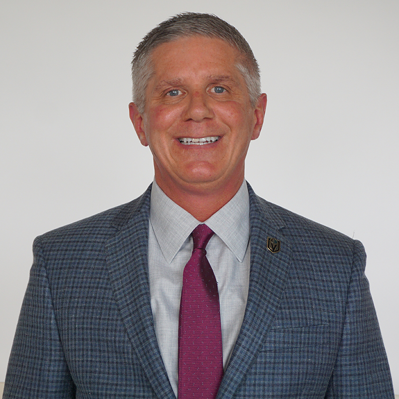 A gray-haired man smiles. He wears business attire with a Vegas Golden Knights pin on his suit jacket.