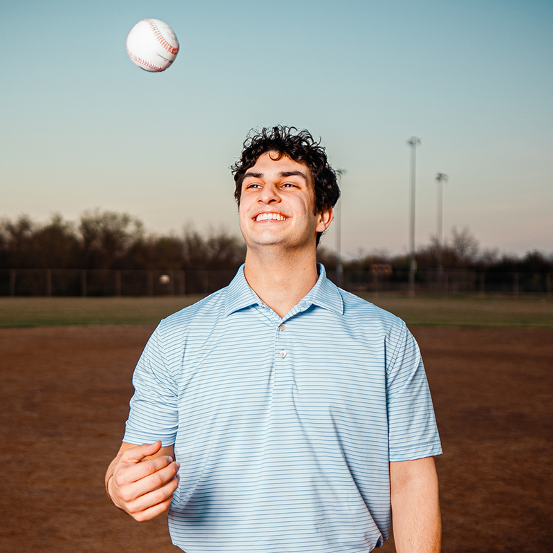 A young man with curly, black hair smiles as he tosses a baseball in the air. He wears a blue polo shirt and stands on a baseball field, representing his interests in sports and business.