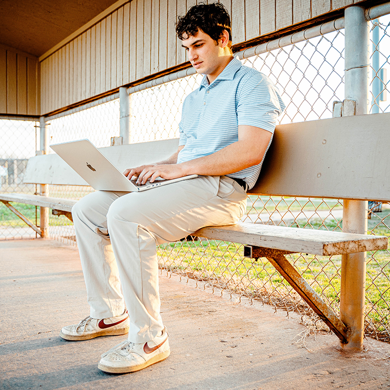 A young man with black, curly hair sits in a baseball dugout and looks at his laptop. He wears a blue polo and khaki pants.