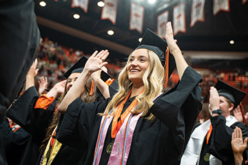 A female student wearing a cap and gown during graduation. She is singing the alma mater and doing the OSU hand sign.