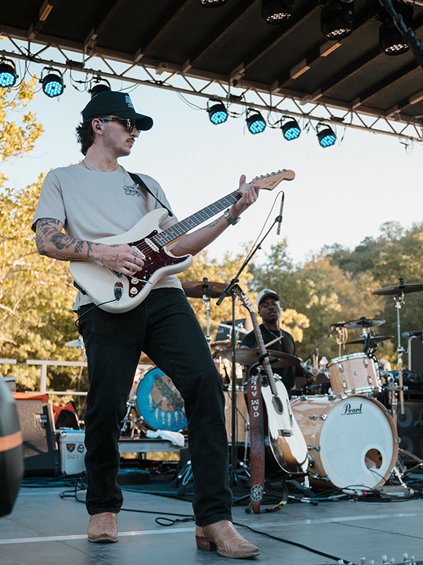 A young man plays electric guitar on a stage. A young man behind him plays drums.