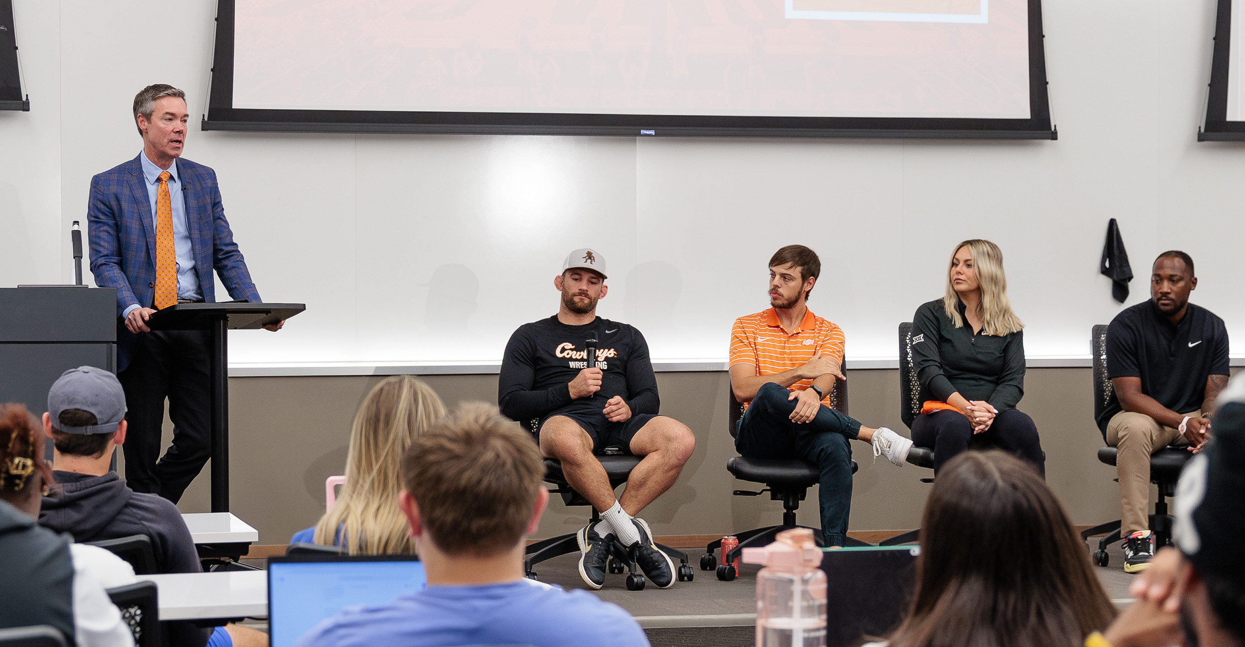 A gray-haired man in a sport coat and orange tie stands at a classroom podium. Four guest speakers, three men and a woman, sit beside him at the front of the room. Students are watching from the classroom rows. The photo is taken from the perspective of the students sitting in the class.