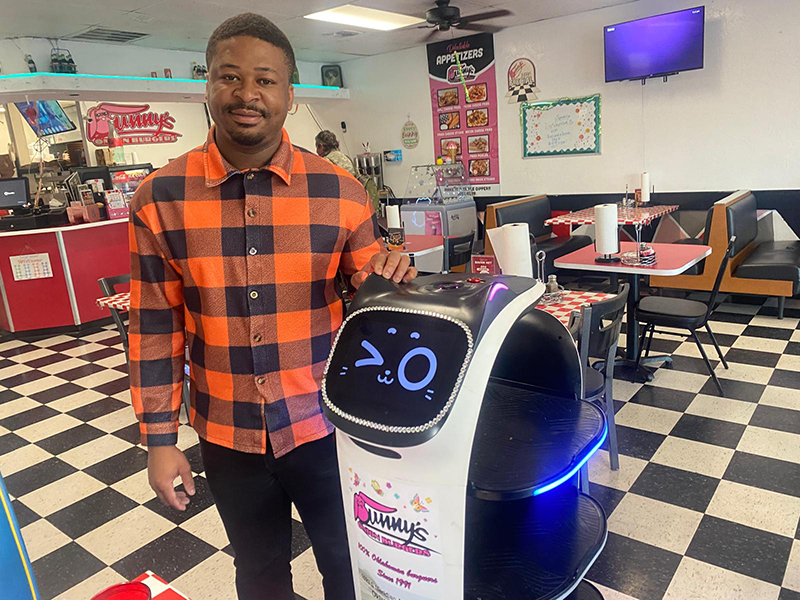 Abraham Terrah stands next to the robot at Bunny's Onion Burgers. The robot has a cute, cartoon cat face.