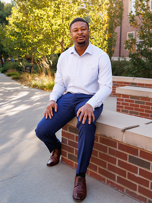 A young man wears business casual attire and dress shoes. He sits on a brick ledge outside a campus building.