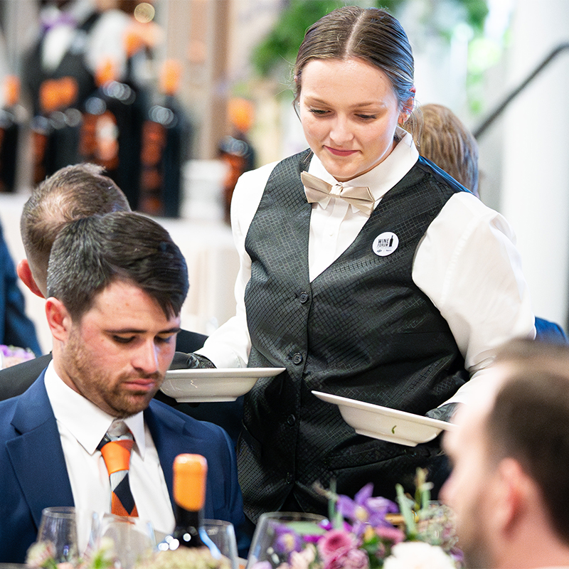 A student waiter in a vest and tie serves the meal to a Wine Forum patron.