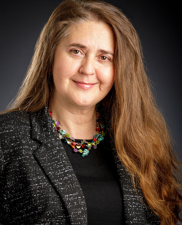 A woman with long hair, dressed in a blazer and adorned with a necklace, exudes professionalism and elegance.