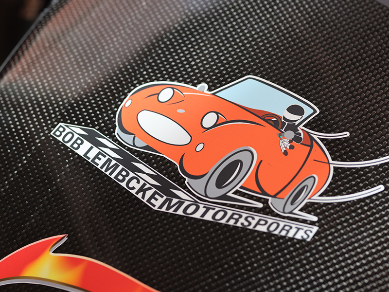 A decal on a carbon fiber racecar that shows an orange race car with feathers and the name Bob Lembcke.
