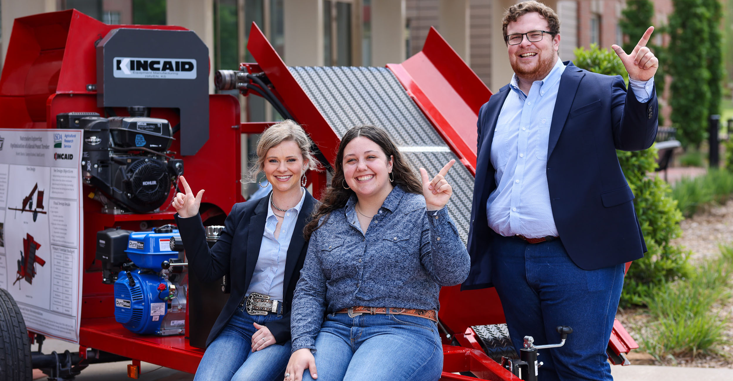 Three students dressed professionally pose in front of their capstone project. A large red machine that assists with agriculture.