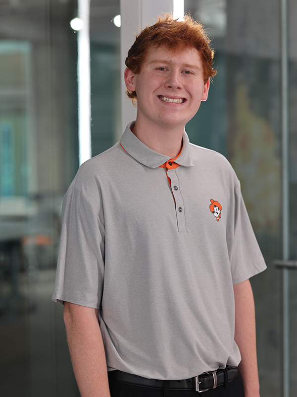 A young man dressed in an OSU polo poses in a modern building hallway.