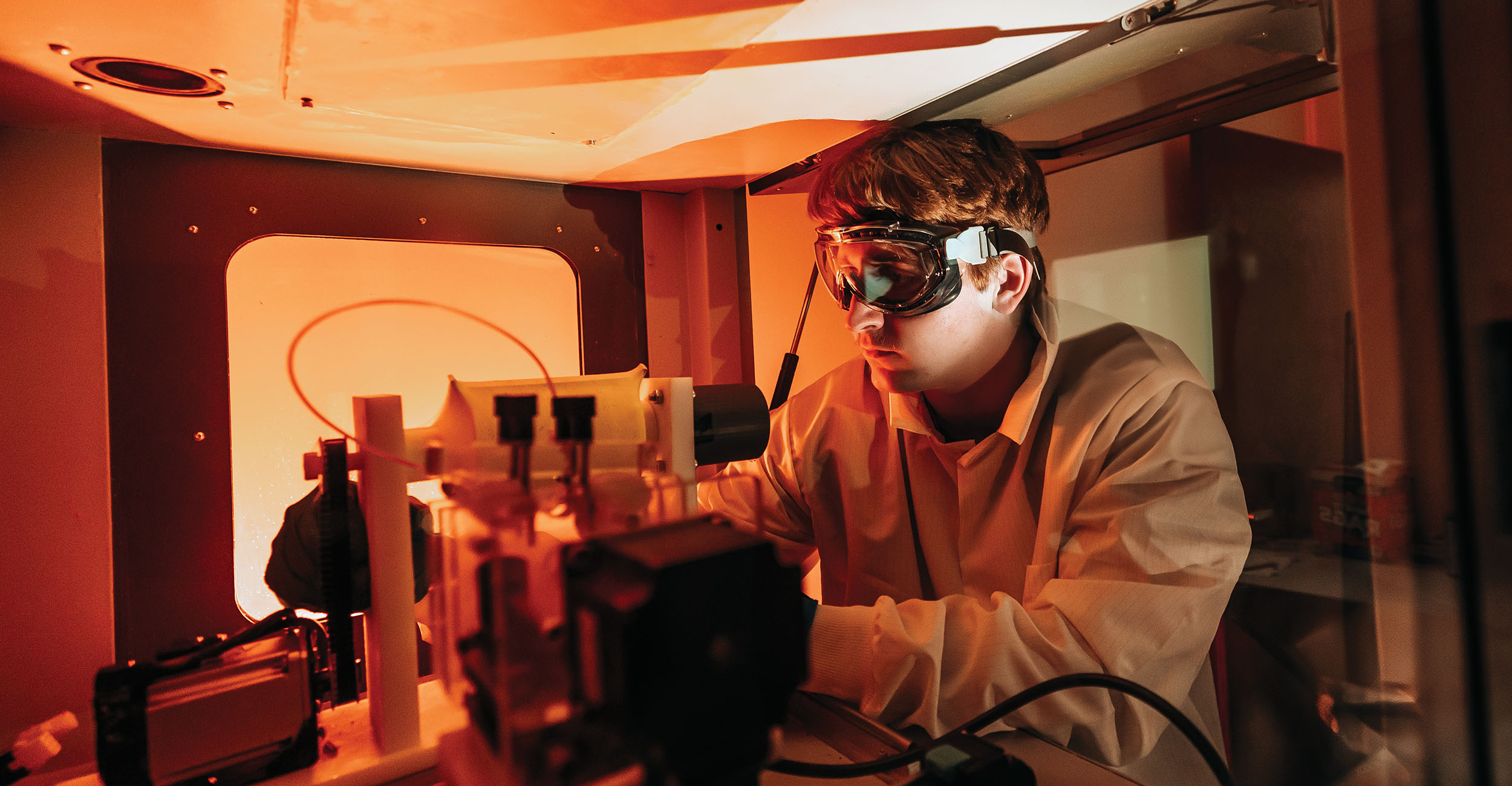 A man in full protective gear works with laboratory equipment in a room that has an orange glow.