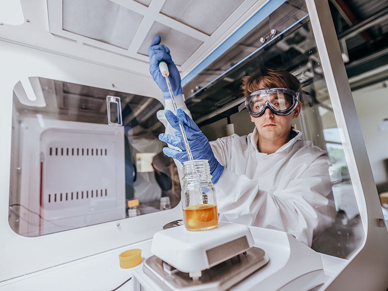 A man dressed in PPE uses a pipet to put liquid in a container within a piece of lab equipment.