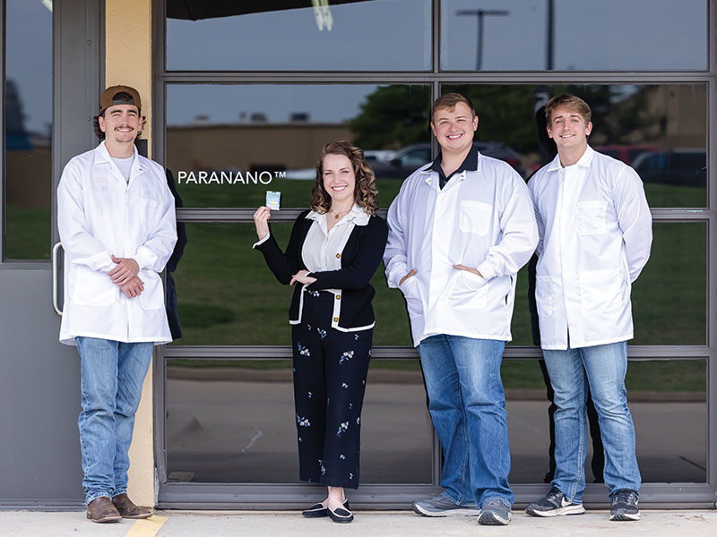 Four people stand outside a building that says, PARANANO. Three men are in white lab coats and one female is holding a package, dressed in business casual attire.