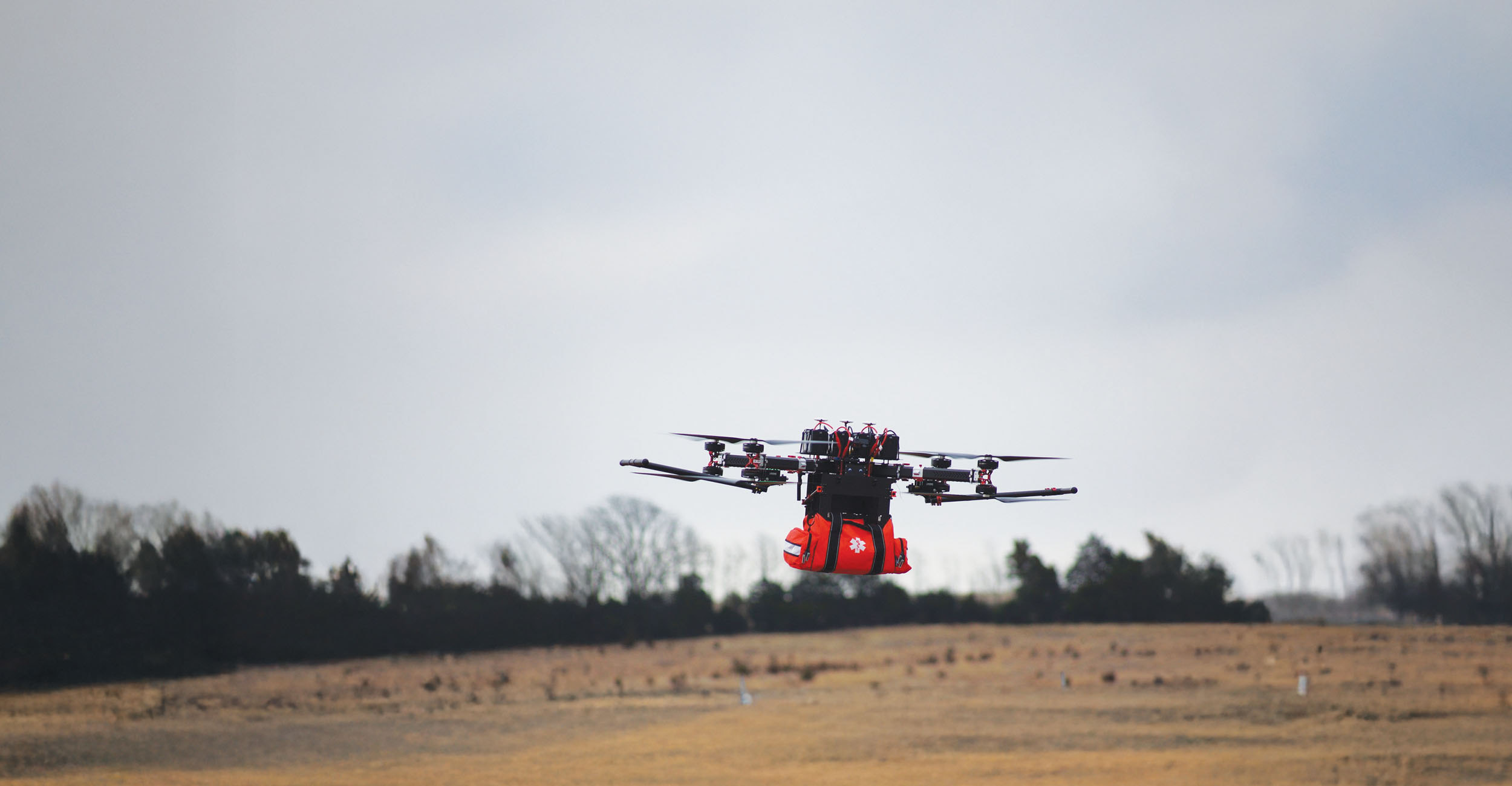 A large drone carrying a medical equipment bag with flat barren land in the background.