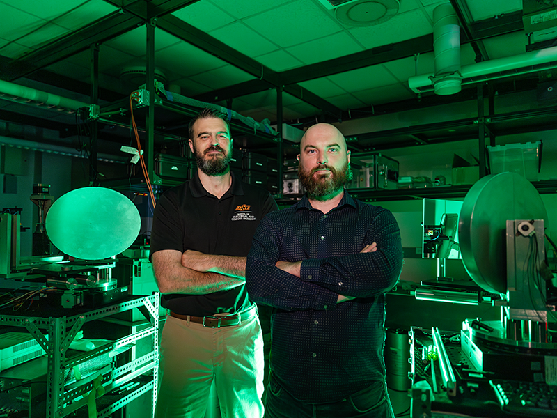 Two men stand inside an engineering laboratory that is illuminated with a green light.