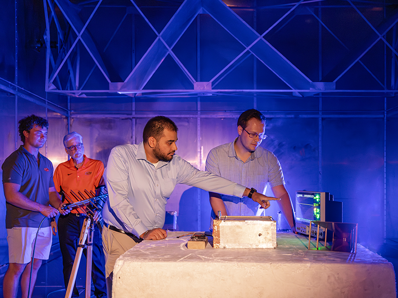 Four men stand inside a steel room inspecting scientific equipment.