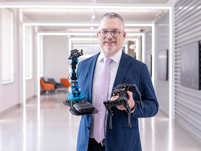 A man in a suit holds robots standing in a modern well lit hallway.