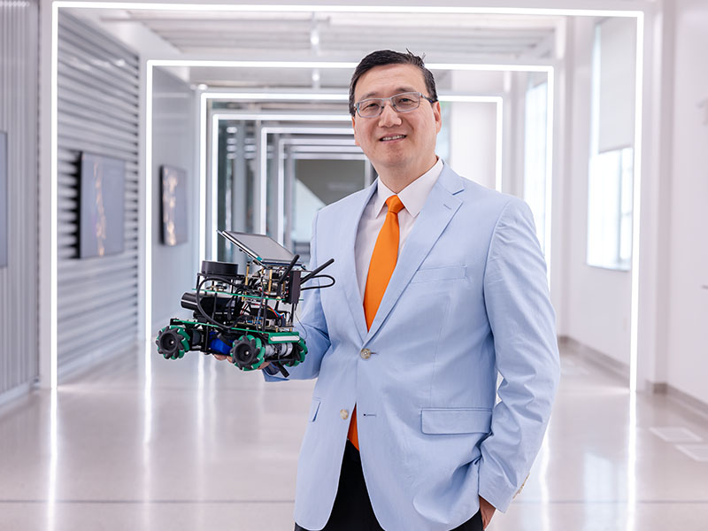 A man in a baby blue suit jacket and orange tie stands in a modern hallway holding a robot.