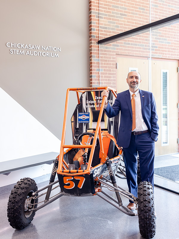 A man in a navy blue suit with orange tie poses with a SAE racecar.