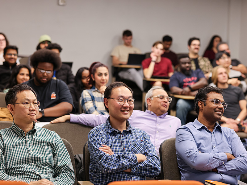 A group of professors sits in the audience of a classroom listening to a presentation, smiling.
