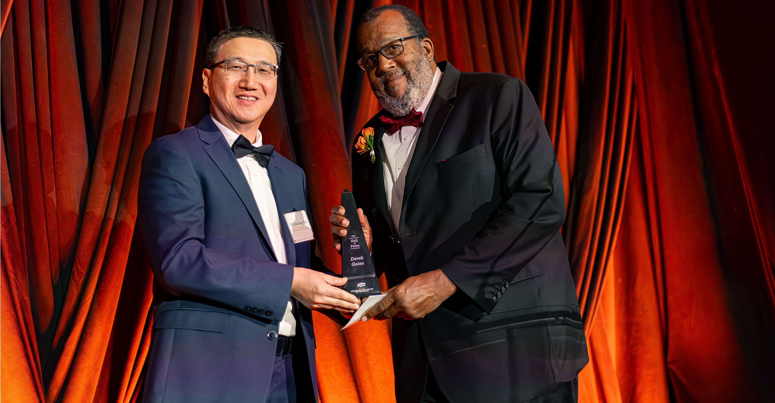 Two people dressed in formal wear stand on an illuminated stage while one individual presents the other with an award.