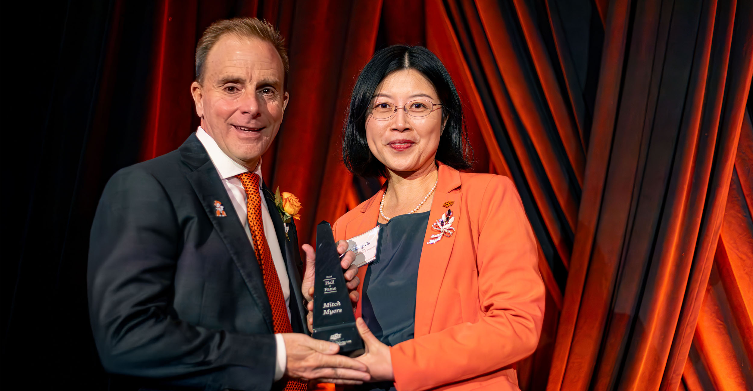 Two individuals stand on an orange illuminated stage while one is awarding the other a certificate.