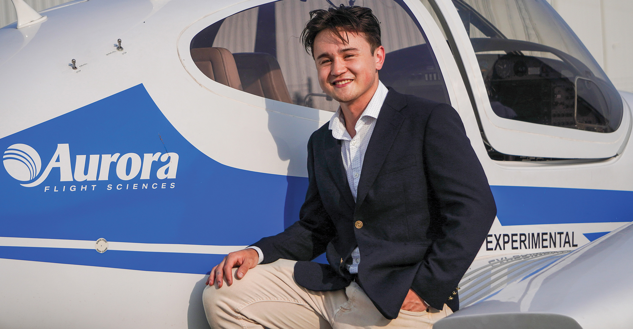A man dressed in business attire poses against the wing of an airplane.